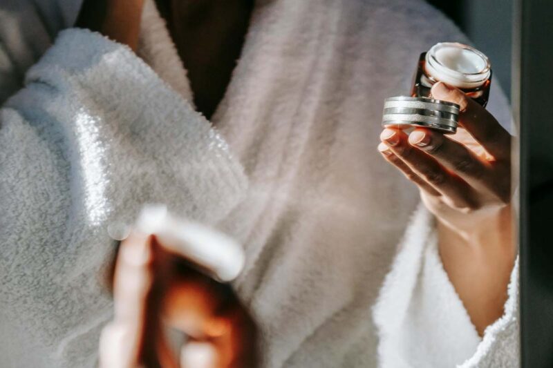 A person wearing a white bathrobe holds a small jar of cream in one hand while preparing to apply it with the other.