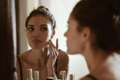 A woman standing in front of the mirror with various skin care products visible on the counter.