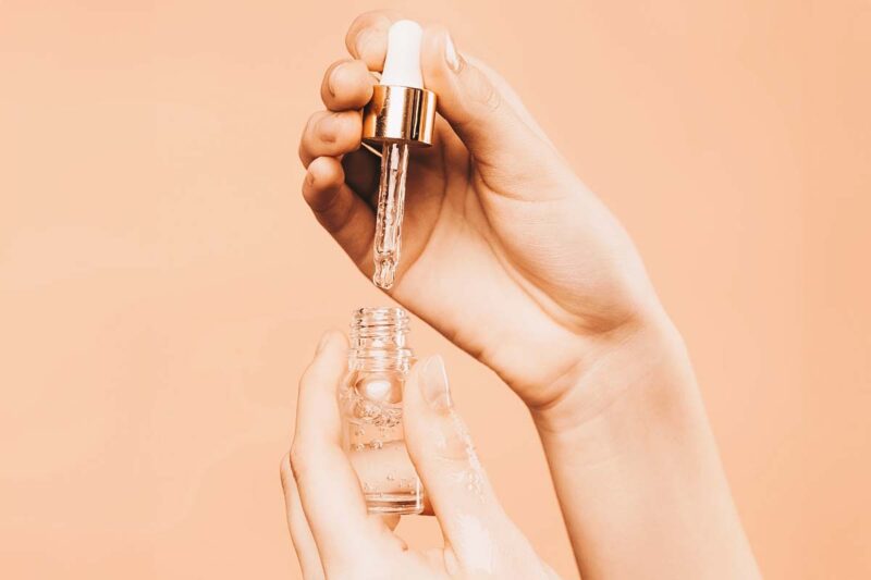 A close-up of two hands using a dropper to extract a liquid from a small glass bottle against a peach-colored background.